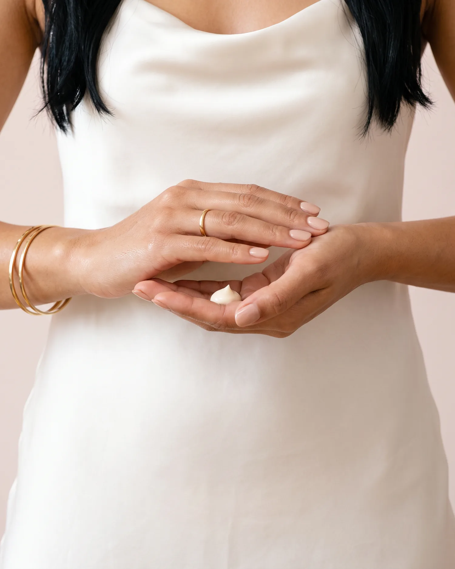 Hands with gold bangles warming a pearl of ivory cream between the palms
