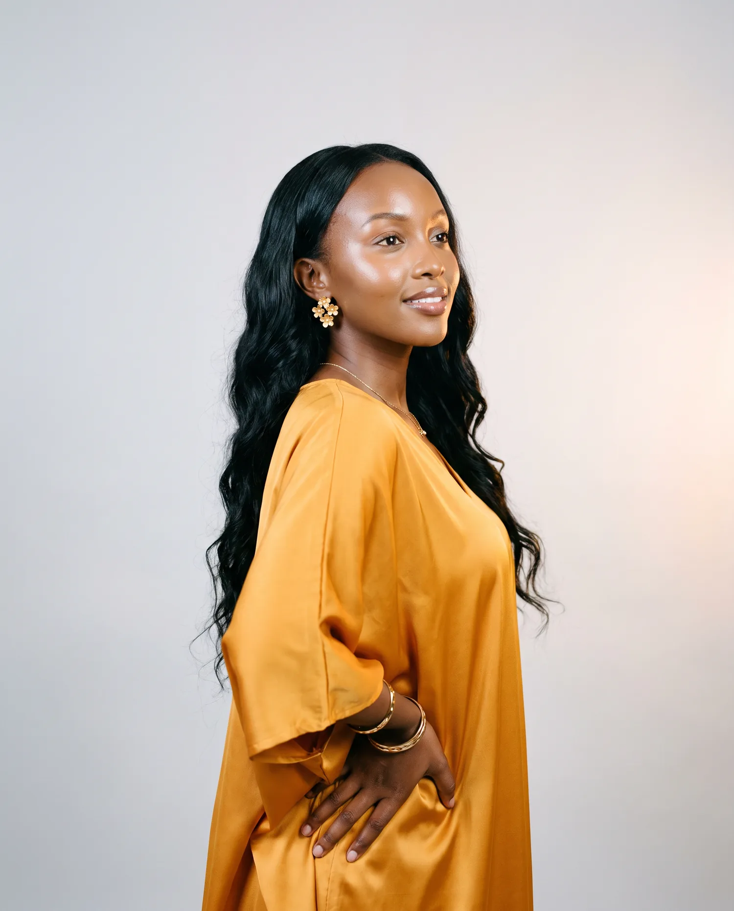 A woman with glowing skin and long dark waves in a marigold satin kaftan turns towards warm light