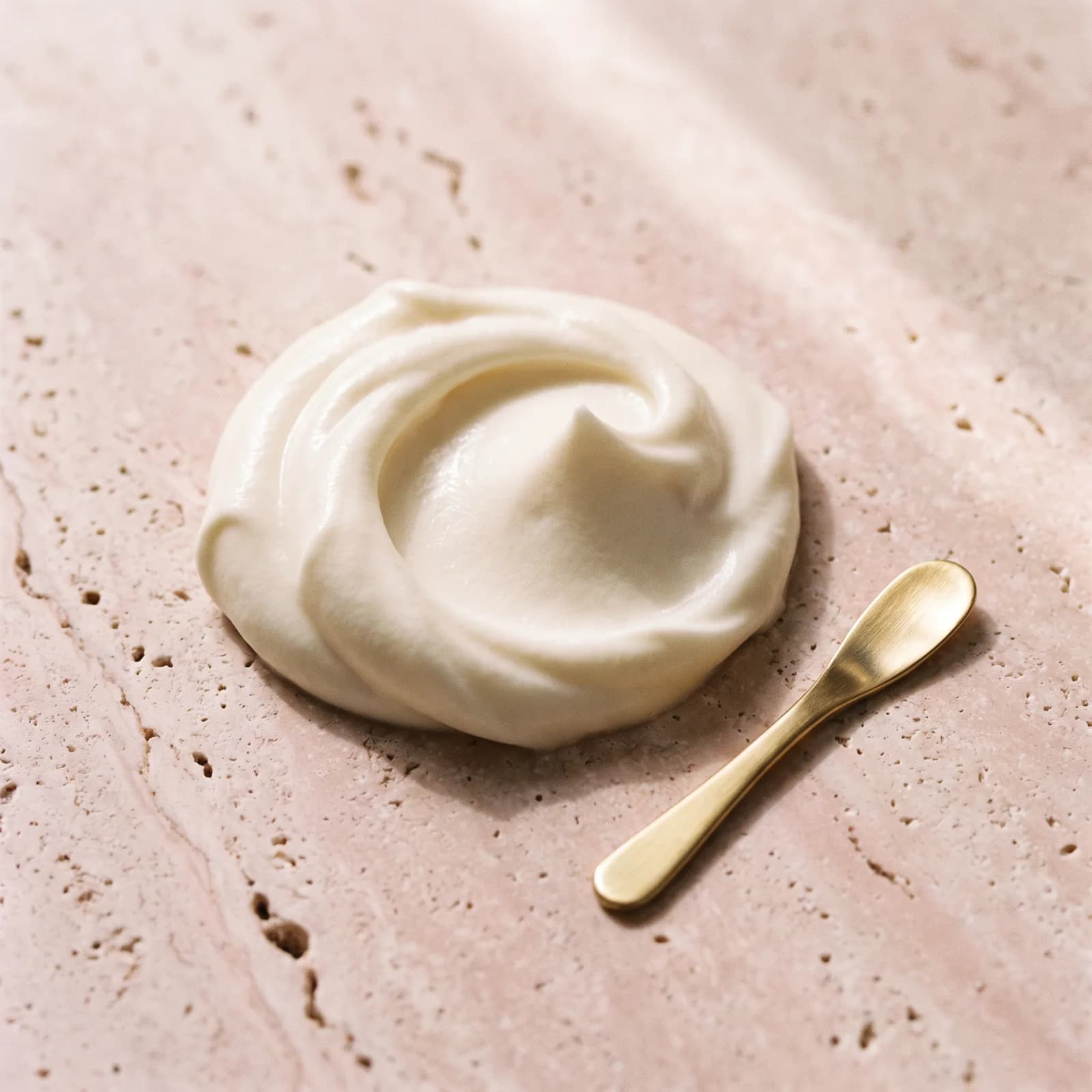 A soft swirl of ivory cream on pink travertine beside a small gold spatula