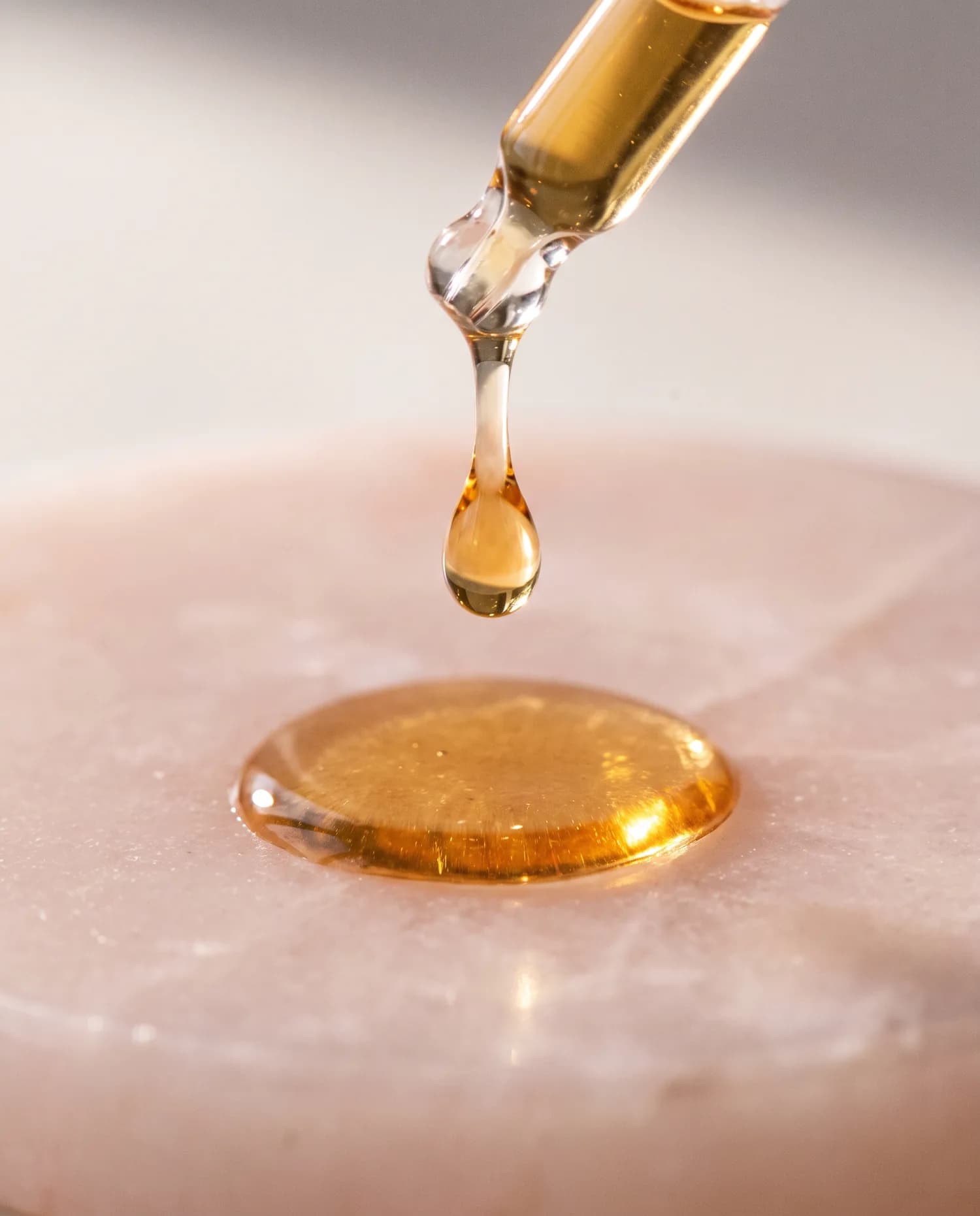 A golden serum drop falling from a glass pipette onto pale pink stone